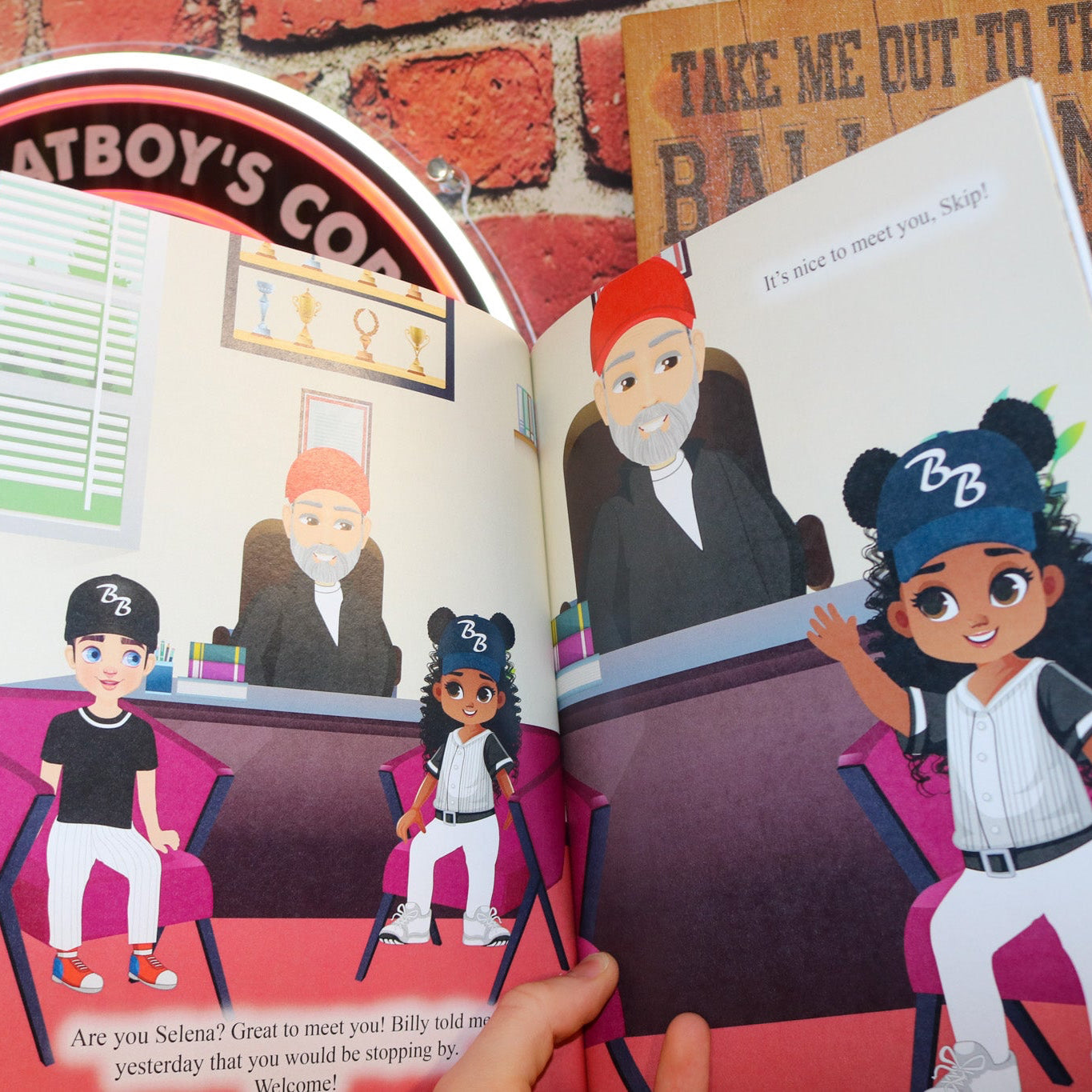 Open book showing illustrations of characters in a baseball-themed setting with a brick wall background.